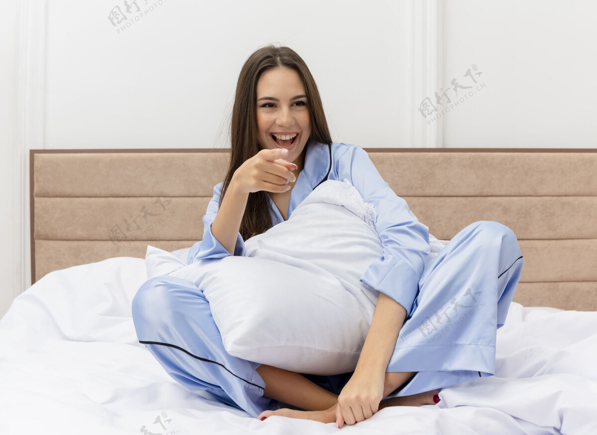手指穿着蓝色睡衣的年轻美女坐在床上 枕着枕头 愉快地微笑着 食指指着卧室内部的摄像头漂亮室内枕头