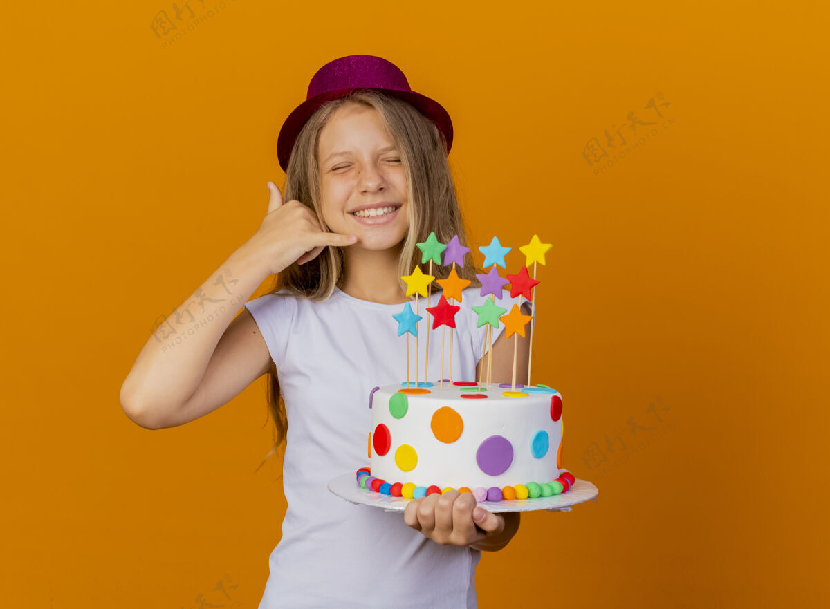 漂亮戴节日帽的漂亮小女孩拿着生日蛋糕微笑着打电话给我的手势 生日派对的概念站着节日手势
