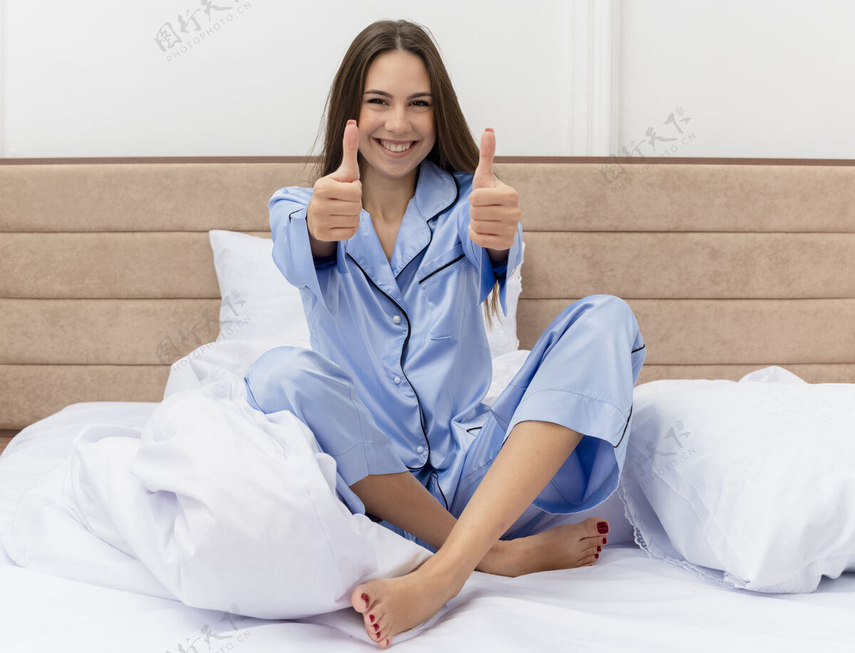 拇指穿着蓝色睡衣的年轻漂亮女人坐在床上休息 竖起大拇指 快乐而积极地微笑 在卧室里享受周末起来坐着蓝色