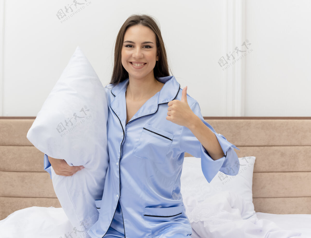 秀穿着蓝色睡衣的年轻美女坐在床上 枕头微笑着 在卧室里竖起大拇指床室内漂亮