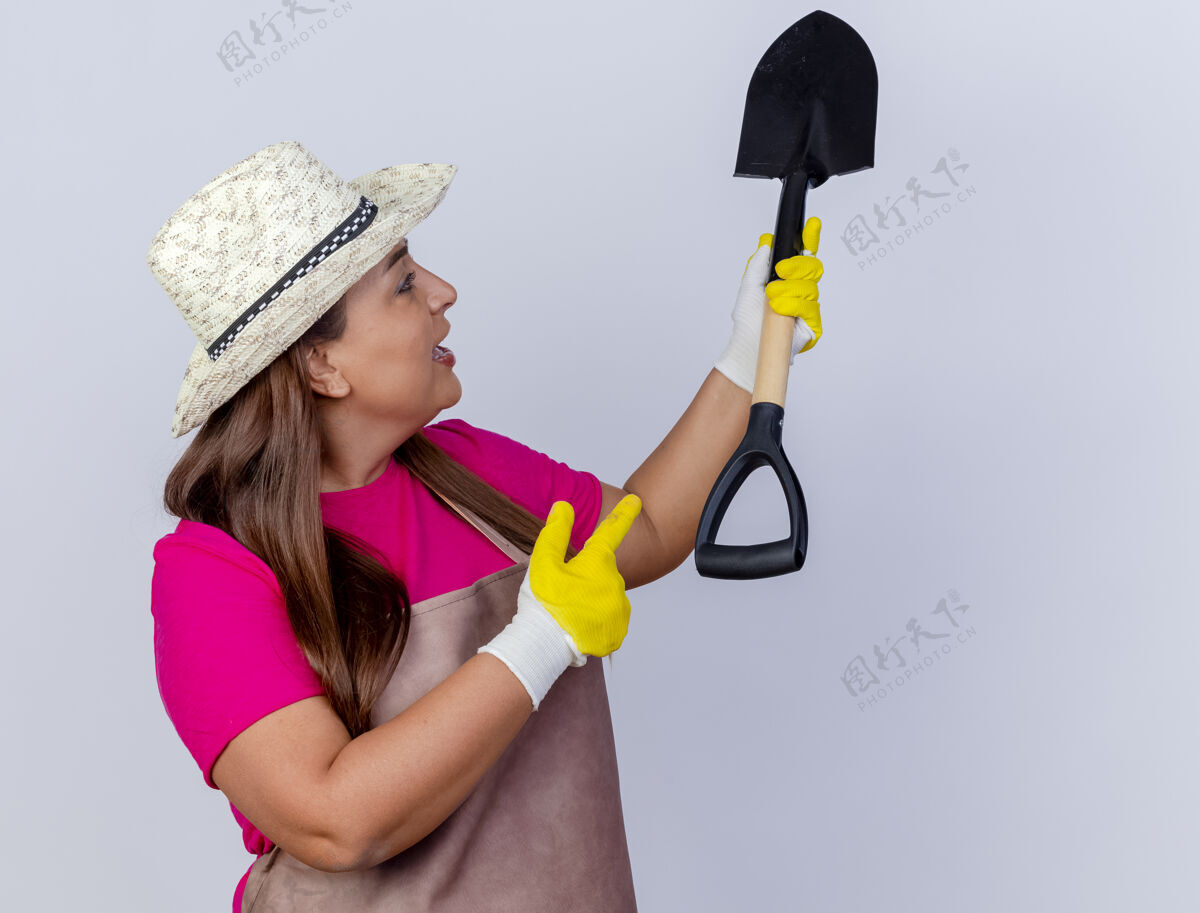花园中年女园丁围着围裙戴着帽子戴着橡胶手套拿着铲子好奇地看着它用手指着它年龄站着女人