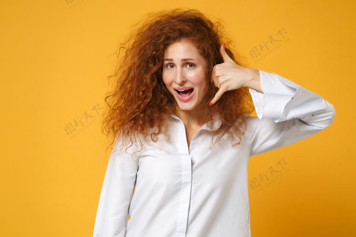 人物兴奋的年轻红发女女孩穿着休闲白衬衫在黄橙色的墙上摆出孤立的姿势秀手势衬衫
