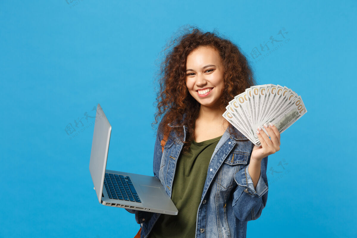 购买年轻的非洲裔美国女孩十几岁的学生在牛仔服装 背包举行个人电脑 球迷的现金钱隔离在蓝色的墙壁上购物使用银行