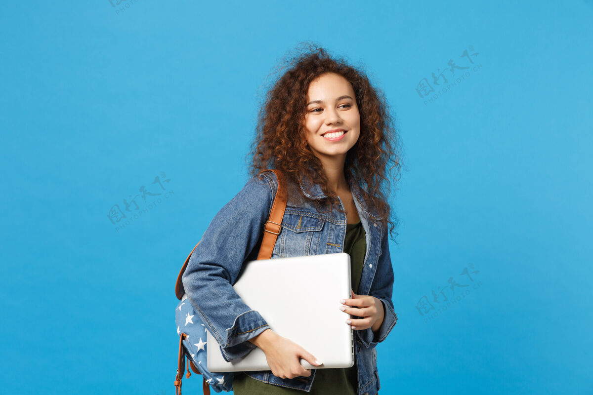 站着年轻的非洲裔美国女孩十几岁的学生在牛仔服装 背包举行电脑隔离在蓝色的墙壁上包笔记本笔记本电脑