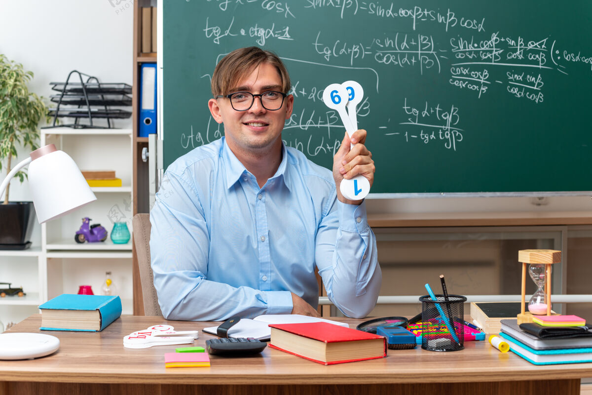 穿着年轻的男老师戴着眼镜 展示着解释课的车牌 微笑着坐在课桌旁 教室里的黑板前放着书和笔记黑板盘子展示