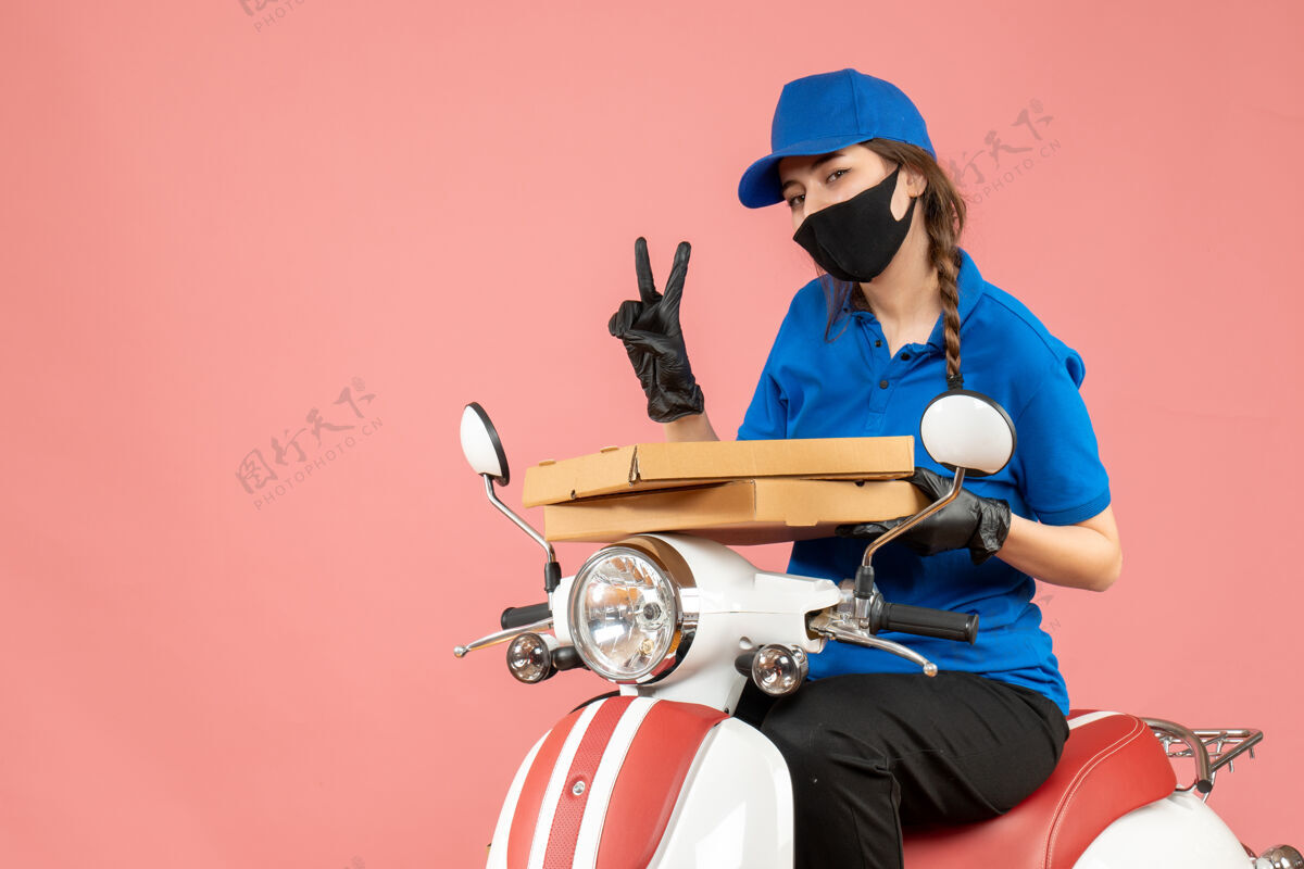 坐俯视图：戴着医用面罩和手套的年轻女快递员坐在滑板车上递送订单 上面有两个粉彩桃手套滑板车面具