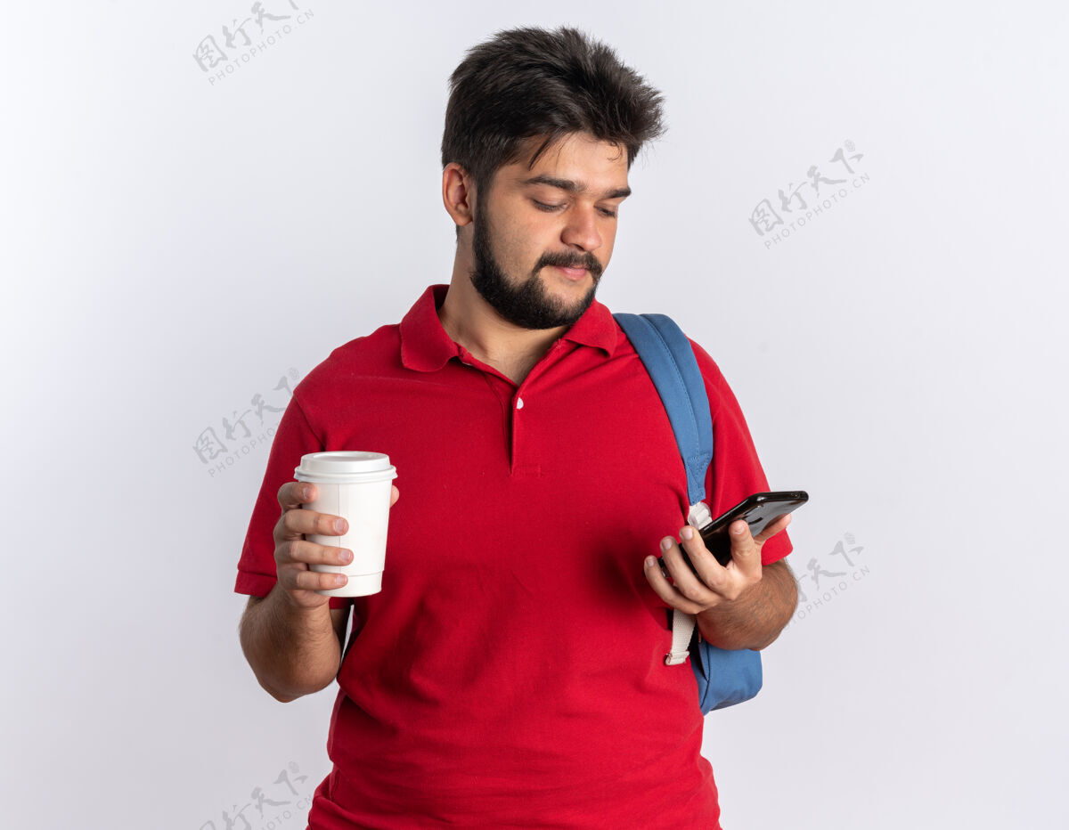 姿势年轻的留着胡子的学生 穿着红色马球衫 背着背包 手里拿着智能手机和咖啡杯 站在白色的墙上 看上去自信地微笑着胡须手势马球
