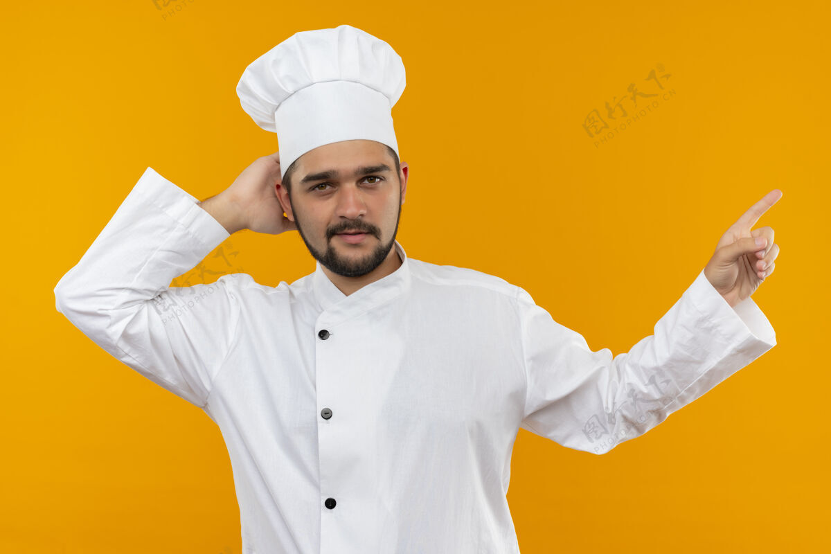 指向身着厨师制服的自信的年轻男厨师把手放在脑后 指着橙色墙上孤立的一面壁板男性手