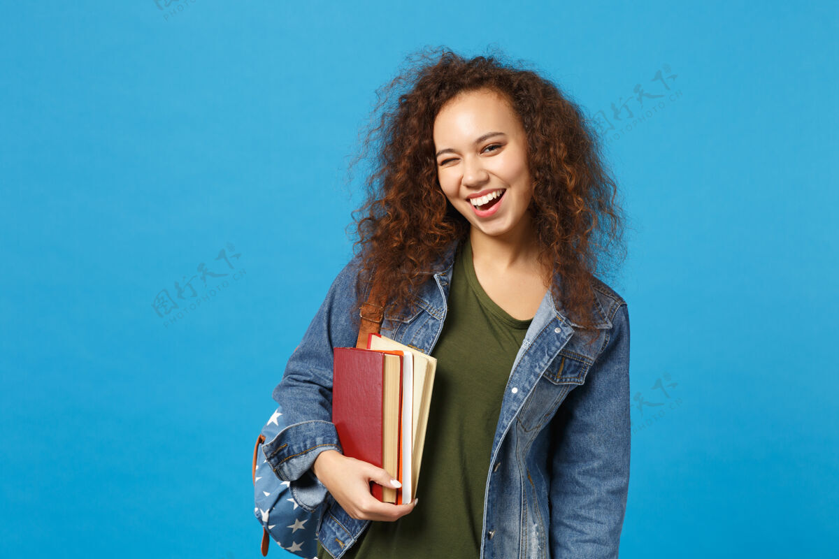 包穿着牛仔服 背着书包的年轻女学生把书孤立地放在蓝色的墙上学校聪明皮肤