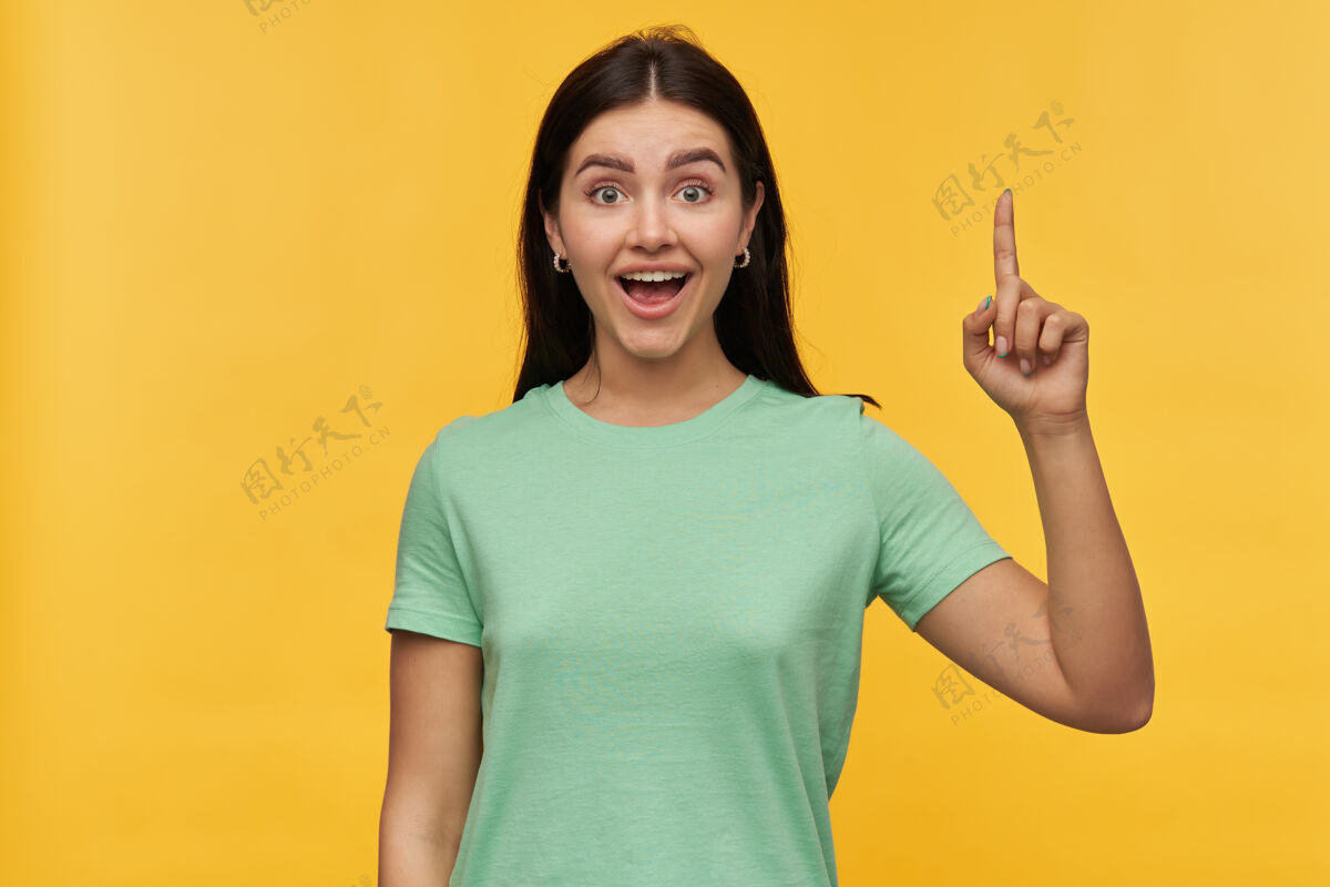 魅力一位身穿薄荷色T恤 留着黑发 心情愉快 兴奋的年轻女子 手里拿着一个伊塞达 用手指着黄色的墙壁惊喜黑发手