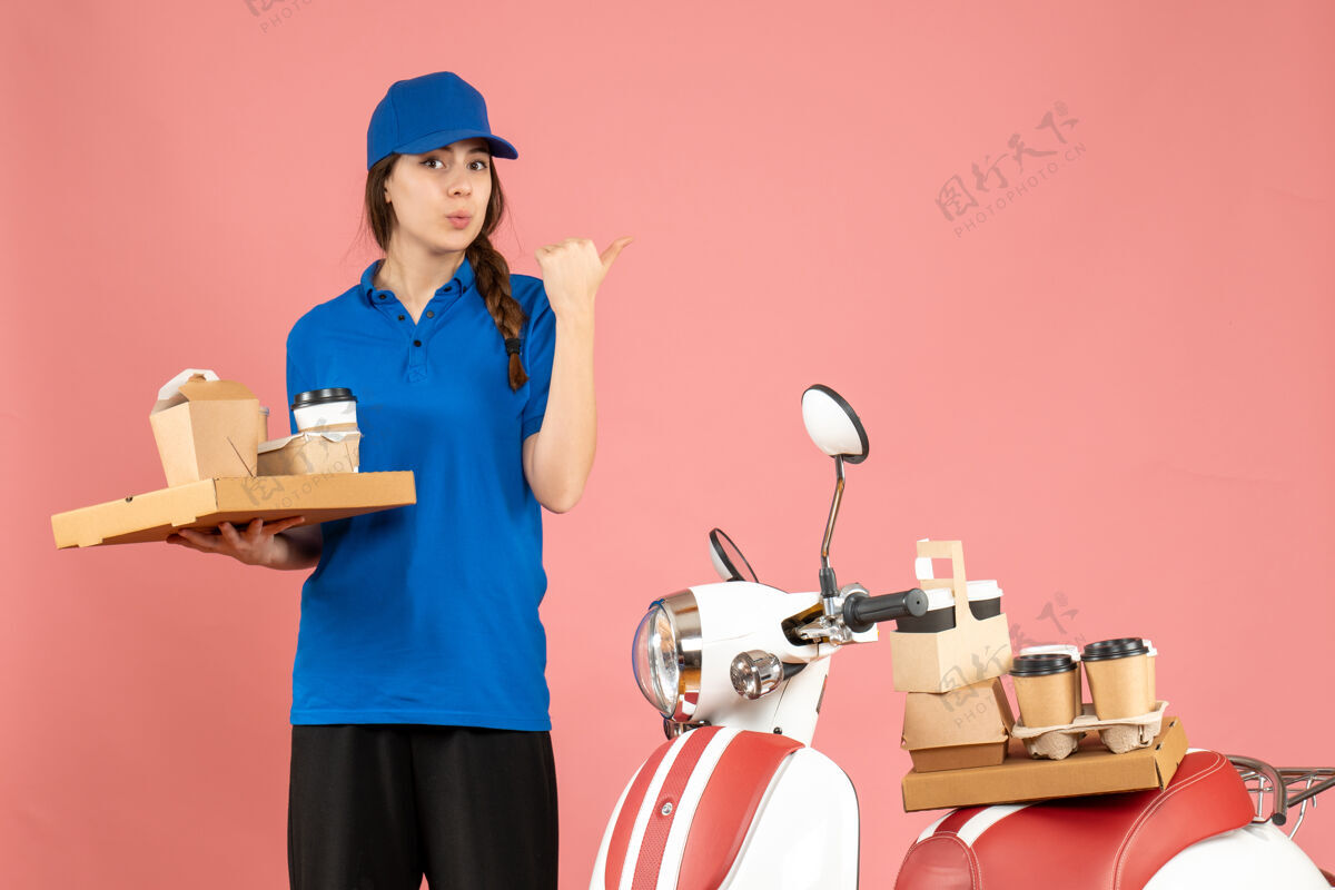 蛋糕前视图的信使女士站在摩托车旁边拿着咖啡和小蛋糕指向粉彩桃色背景背面专业成人微笑