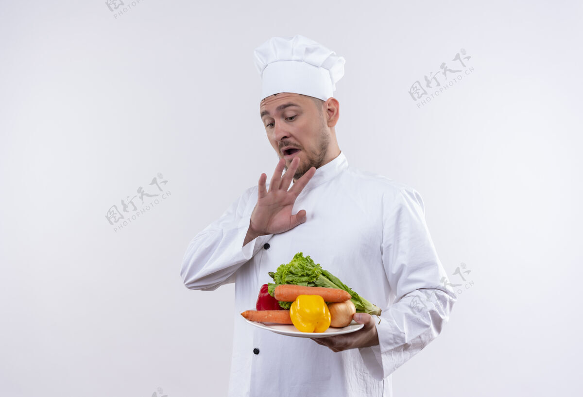 印象年轻帅气的厨师穿着厨师制服 手里拿着盛有蔬菜的盘子 手放在上面 放在隔离的白色墙壁上 留着复印空间烹饪持有制服