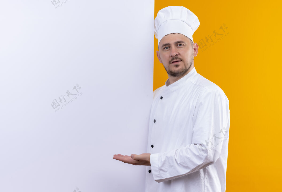 自信自信的年轻帅哥厨师身着厨师制服站在白色的墙后 橙色的墙上有一个单独的复制空间烹饪帅气站立