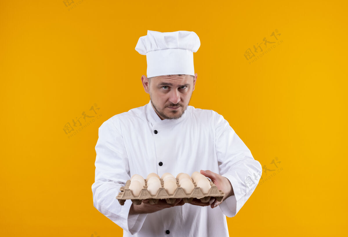厨师自信的年轻帅哥厨师身着厨师制服 手里拿着一盒鸡蛋 隔离在橙色的墙上制服厨艺纸箱