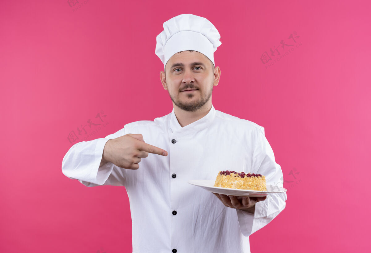 盘子自信的年轻帅哥厨师身着厨师制服 拿着一盘蛋糕指着隔离在粉红色墙上的蛋糕制服自信指着