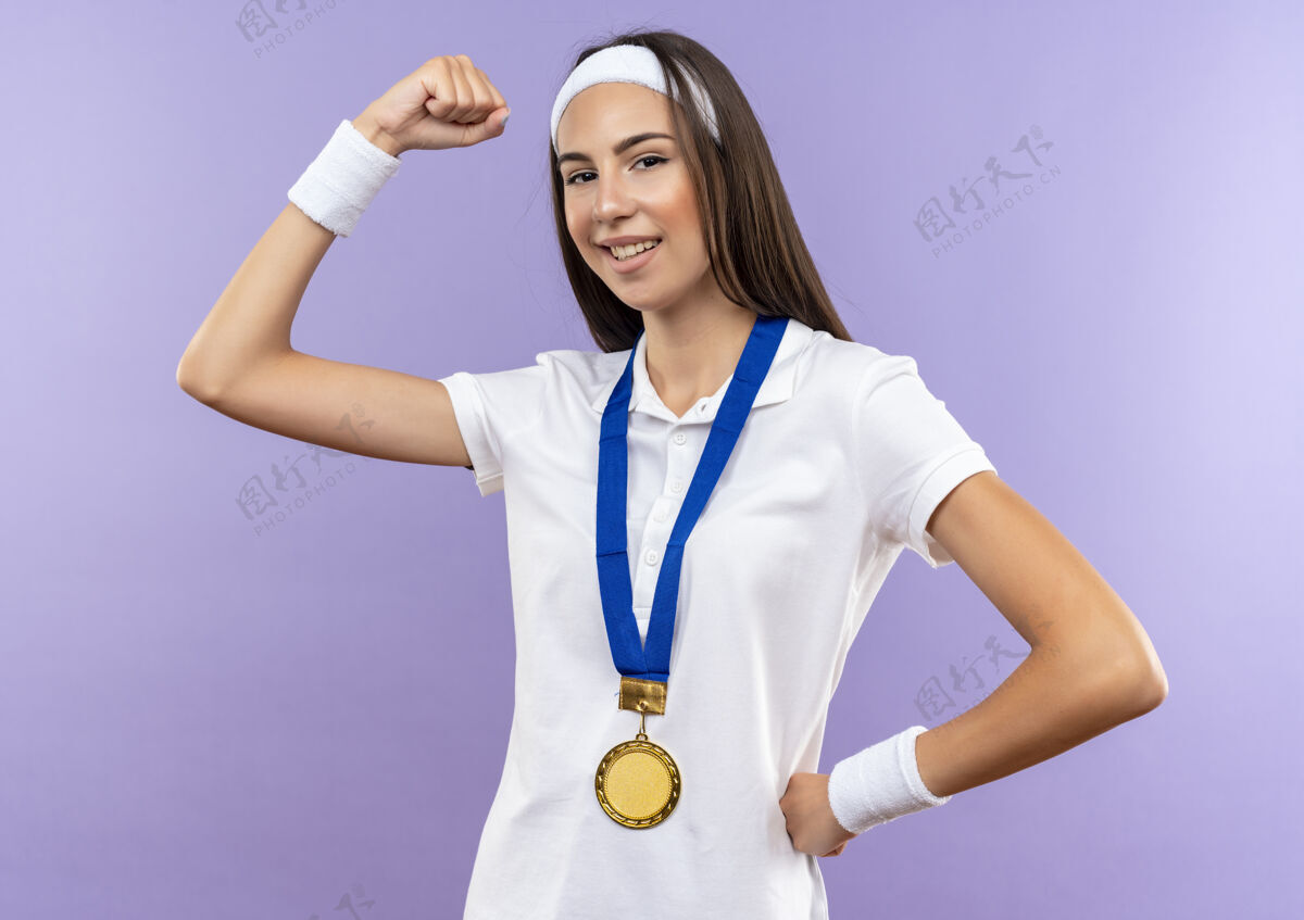 女孩自信漂亮的运动女孩戴着头带和腕带和奖牌 手放在腰上 在紫色的墙上孤立的强有力的手势腕带强壮腰