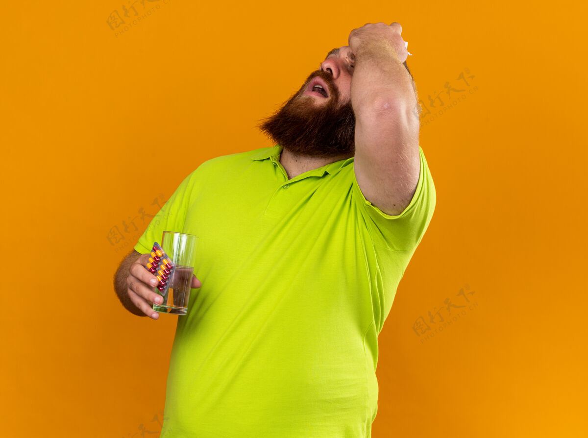 抱着穿着黄色马球衫 留着不健康胡须的男人 手里拿着一杯水和药丸 站在橘黄色的墙上 感觉冷得发烧 头痛得厉害男人药丸胡子
