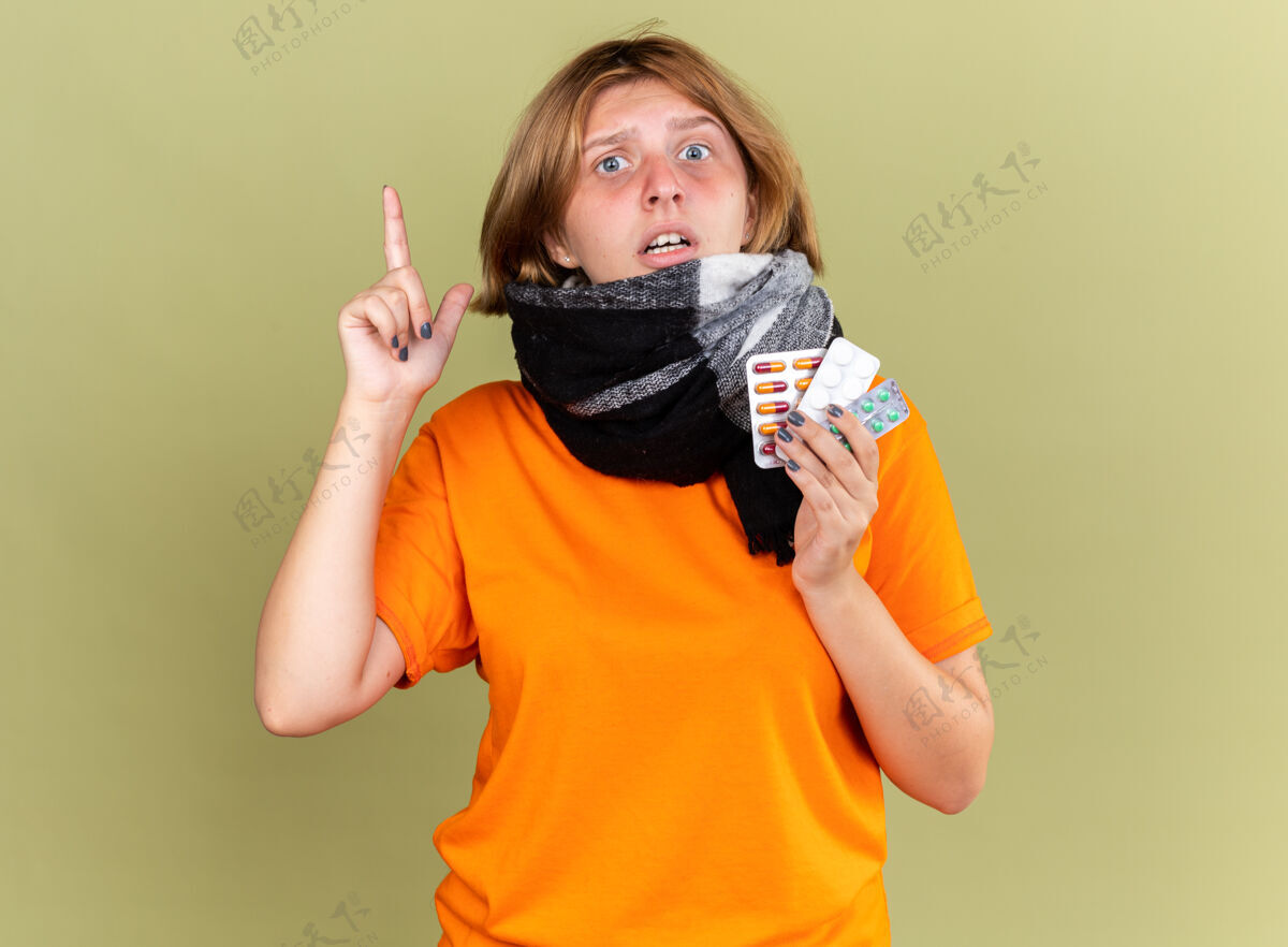 女人不健康的年轻女子穿着橙色t恤 脖子上围着暖和的围巾 感觉不舒服 患流感 拿着不同的药片 看起来很困惑 食指站在绿色的墙上围巾不同感觉