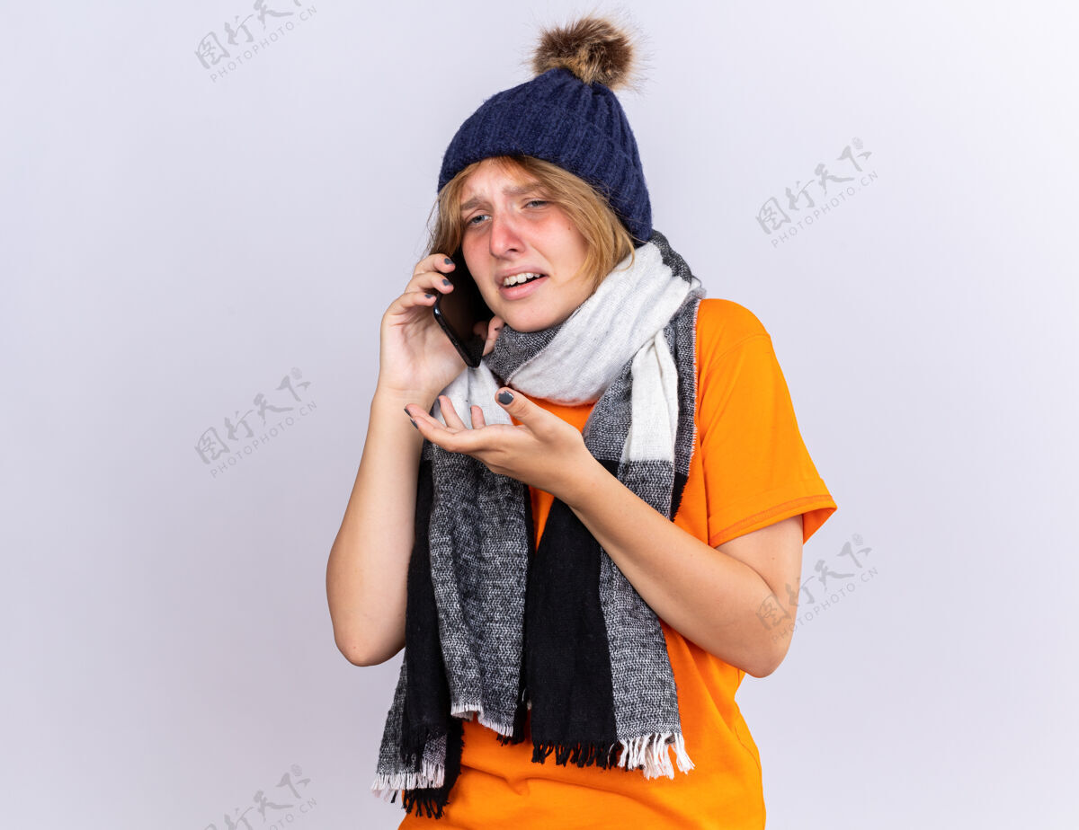 痛苦不健康的年轻女子穿着橙色t恤 脖子上围着暖和的围巾 戴着帽子 感觉很糟糕 患了流感 在手机上说 表情悲伤 站在白色的墙壁上担心女人围巾表情