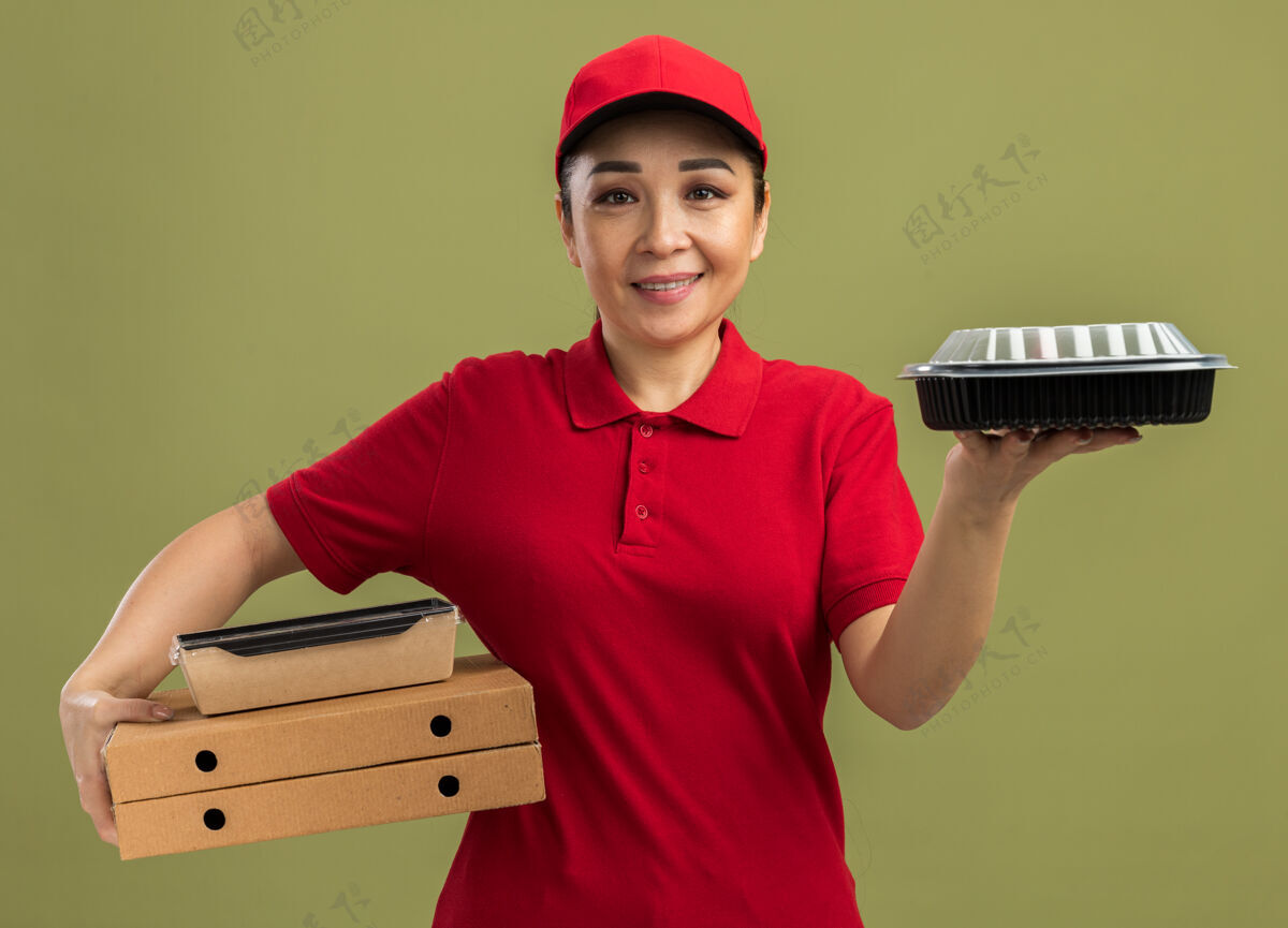 拿着身穿红色制服 头戴鸭舌帽的年轻送货员 手拿披萨盒和食品包 脸上带着微笑站在绿色的墙上制服站着披萨