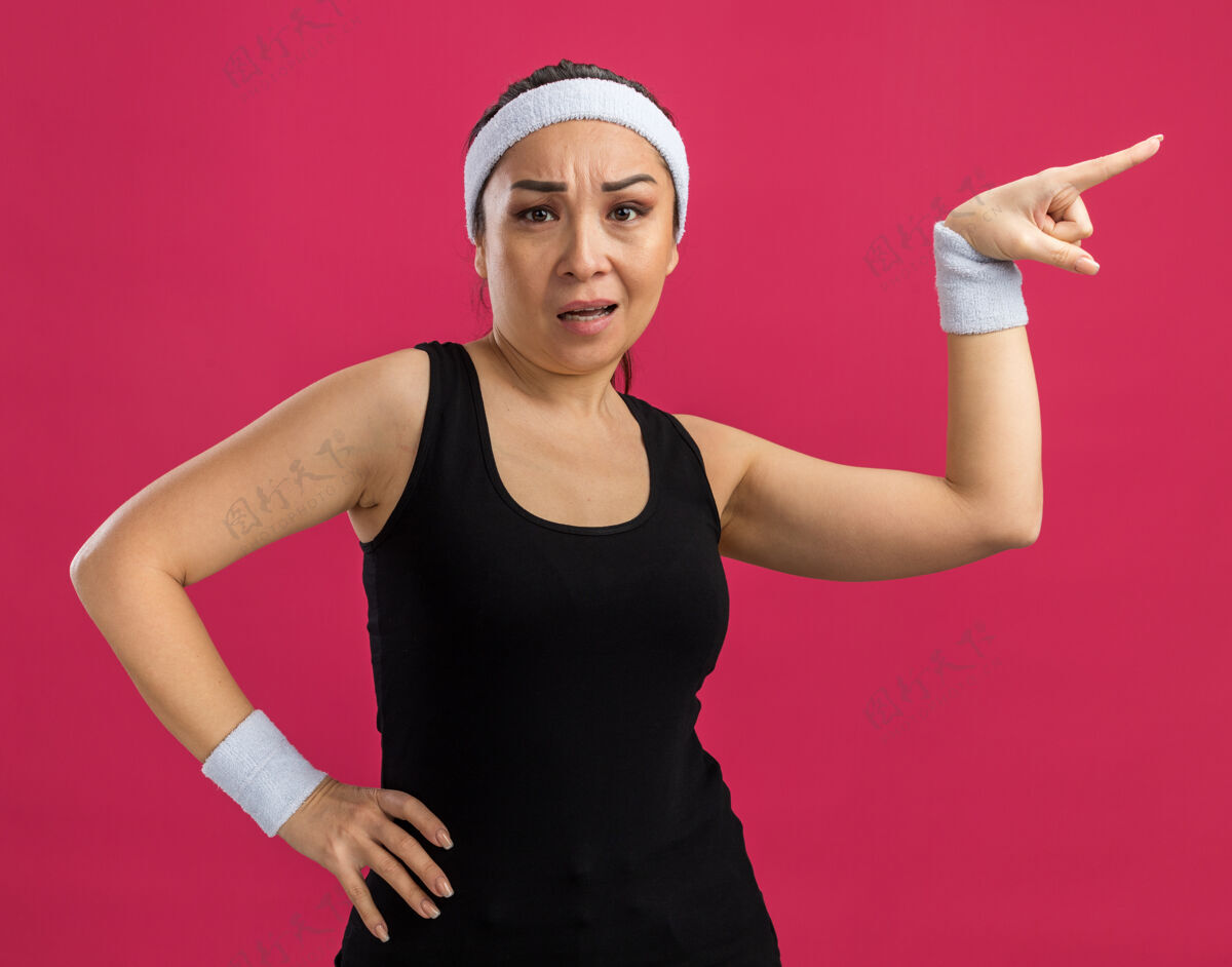 站戴着头巾的年轻健身女士困惑地用食指指着站在粉红色墙上的一边女人壁板困惑