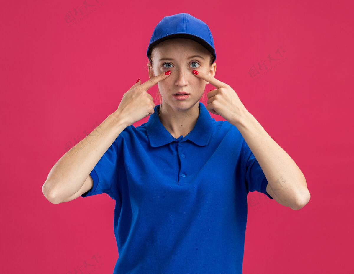 交货身穿蓝色制服 头戴鸭舌帽的年轻送货女 严肃的脸上用食指指着站在粉红色墙上的眼睛严重年轻指向