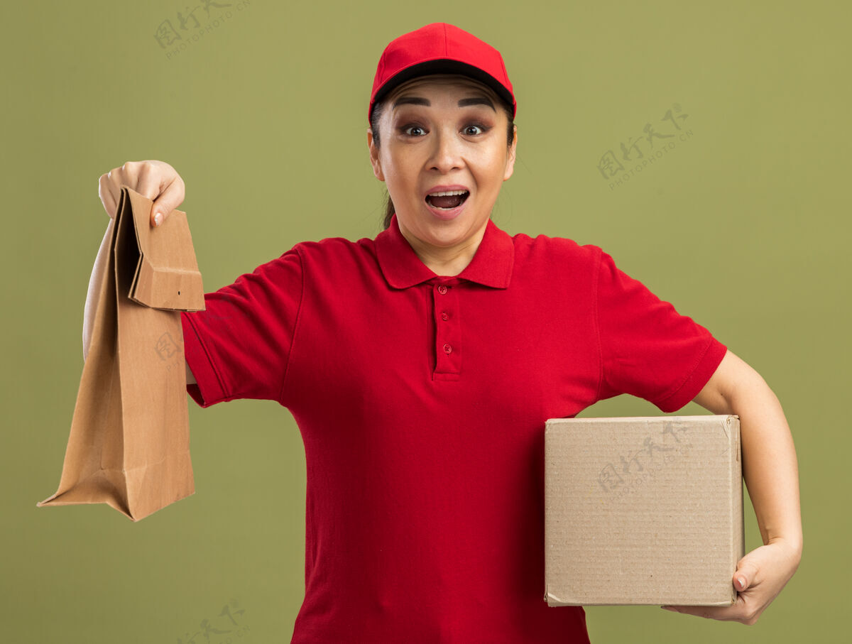女人身穿红色制服 头戴鸭舌帽 手拿纸包和纸箱的年轻送货员站在绿色的墙边 又高兴又惊讶帽子惊喜年轻