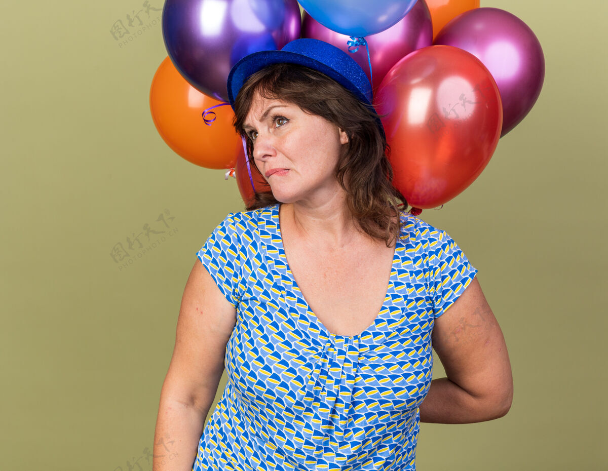 一旁中年妇女戴着派对帽 手里拿着一堆五颜六色的气球 一边看一边歪歪着嘴 带着失望的表情站在绿色的墙上庆祝生日派对表情年龄立场