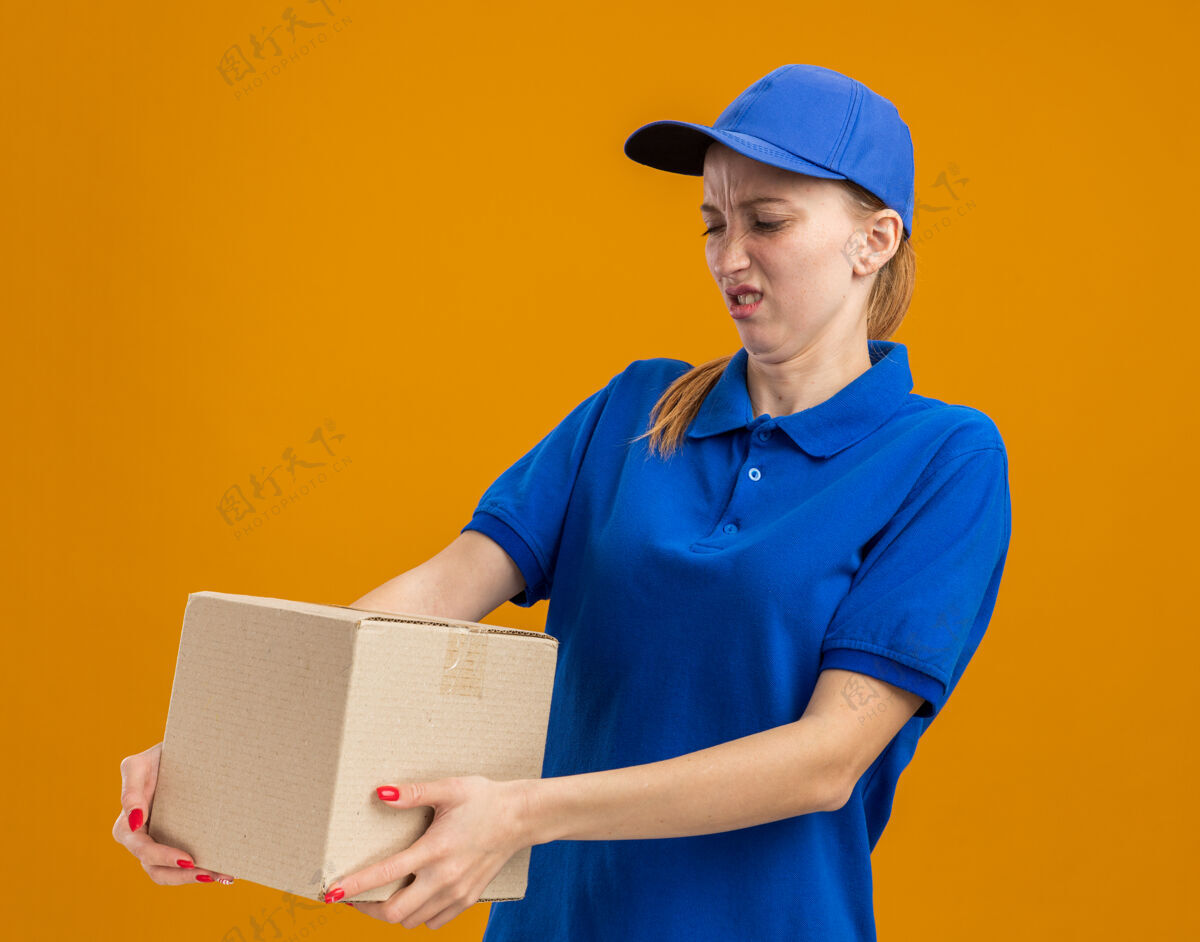 送货穿着蓝色制服 戴着帽子的年轻送货女孩站在橙色的墙上 拿着一个纸板箱 看着它困惑和不高兴制服抱着不高兴