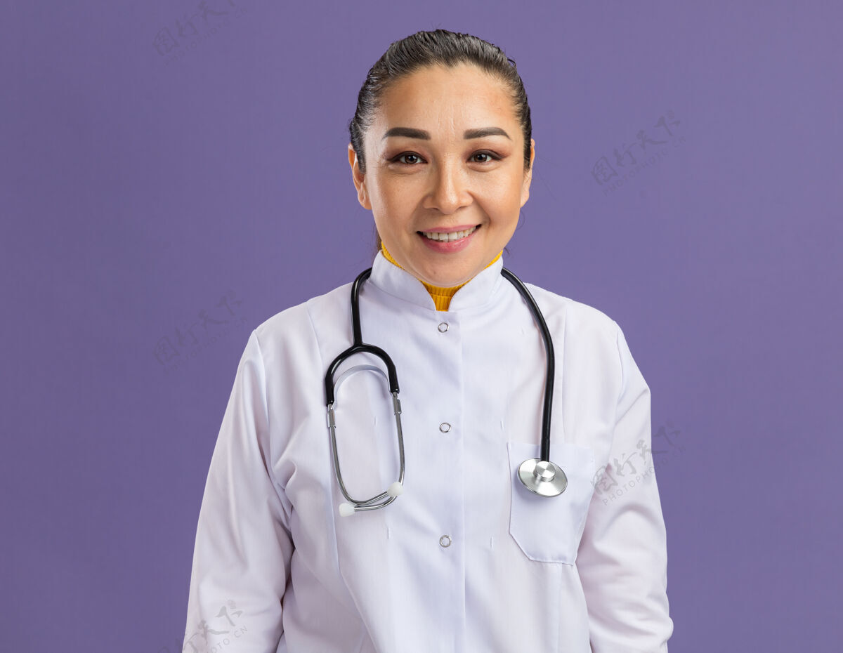 微笑身穿白色药衣 脖子上戴着听诊器的年轻女医生站在紫色的墙上 微笑着自信地站着周围外套站