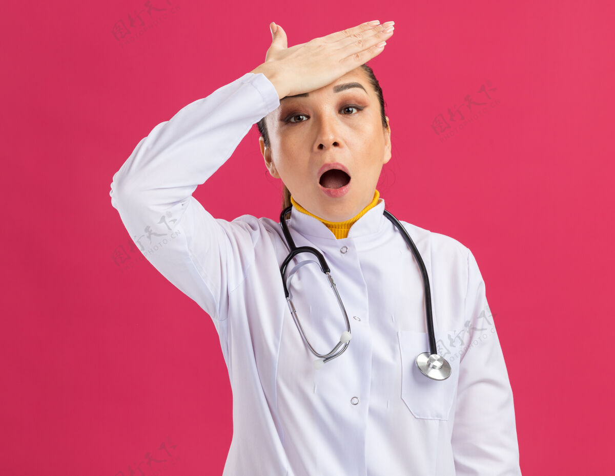 额头年轻的女医生 穿着白药衣 脖子上戴着听诊器 额头上戴着一只手 误站在粉红色的墙上手女人困惑