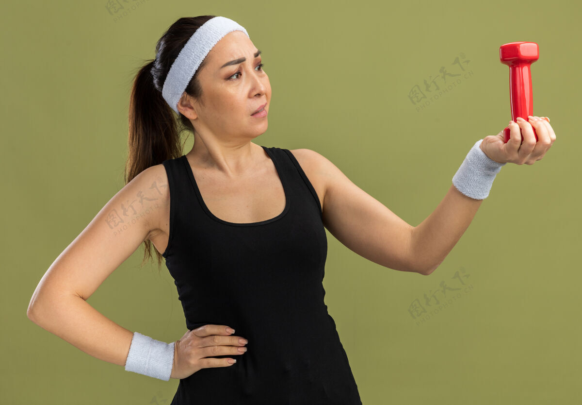 女人年轻的健身女戴着头带 带着哑铃 站在绿色的墙上困惑地看着它站健身头带