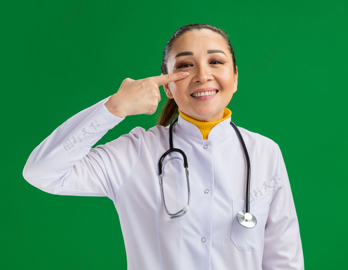 听诊器年轻的女医生 穿着白药衣 脖子上戴着听诊器 用食指指着眼睛 站在绿色的墙上欢快地笑着指着围着手指
