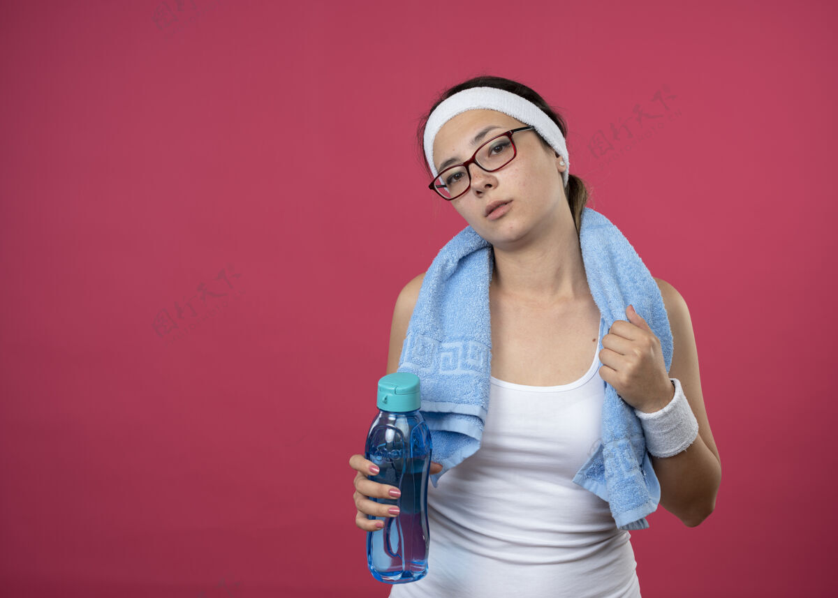 头带自信的年轻运动女孩戴着眼镜 脖子上围着毛巾 戴着头带 手腕上拿着水瓶持有毛巾光学