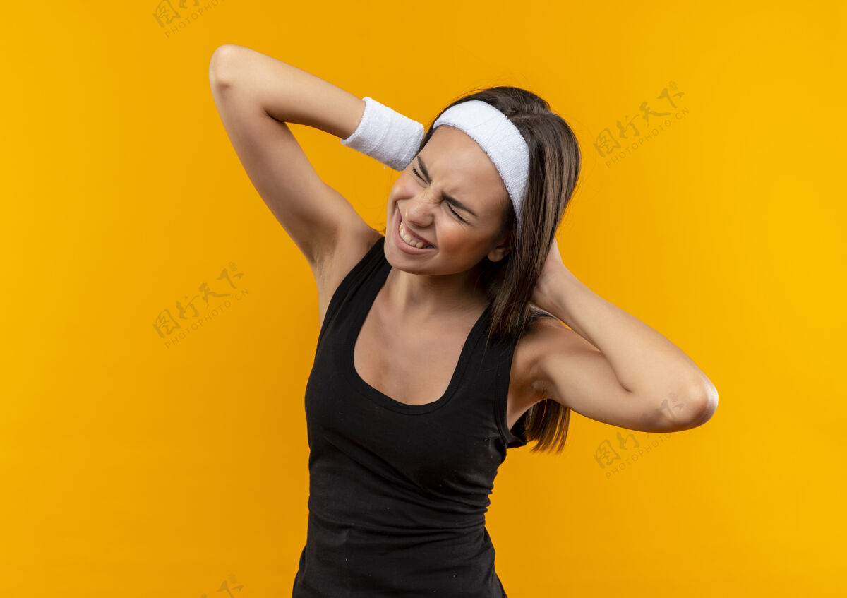 运动疼痛的年轻漂亮的运动女孩戴着头带和腕带把双手放在她的头后面橙色的墙上戴疼头带