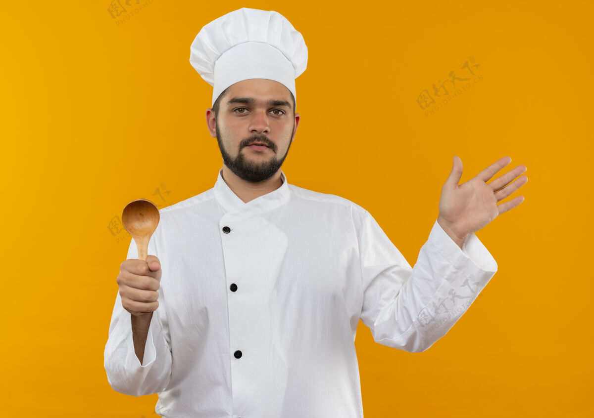 显示自信的年轻男厨师穿着厨师制服 拿着勺子 在橘色的墙上展示着孤立的空手信心手烹饪