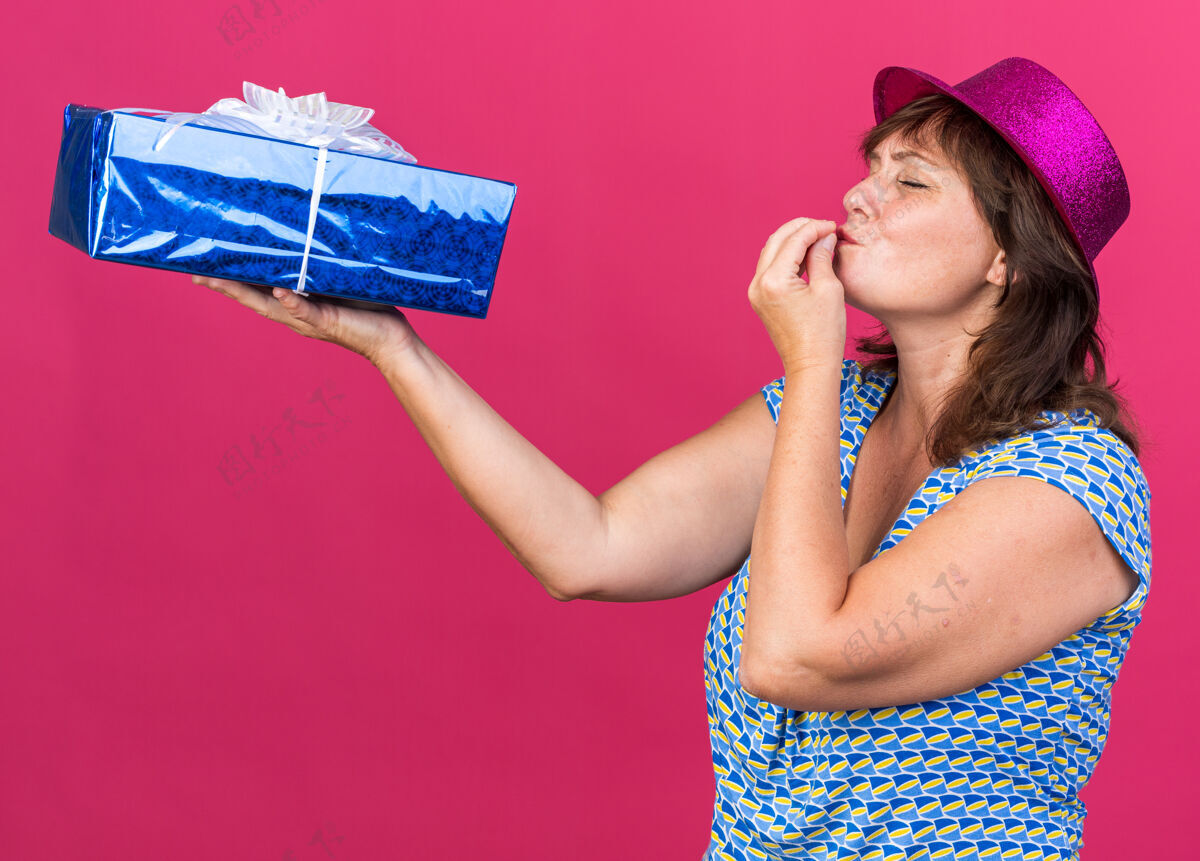 中间中年妇女戴着派对帽拿着礼物看着它做着美味的手势站在粉色的墙上庆祝生日派对手立场庆祝