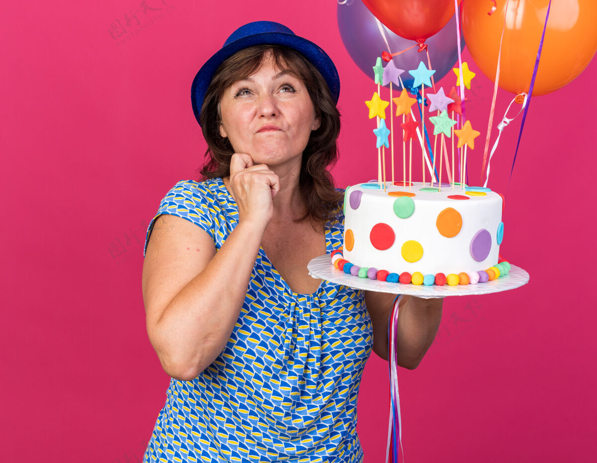 沉思中年妇女戴着派对帽 手里拿着五颜六色的气球 手里拿着生日蛋糕 神情沉思地抬起头 站在粉色的墙上想着庆祝生日派对帽子女人生日