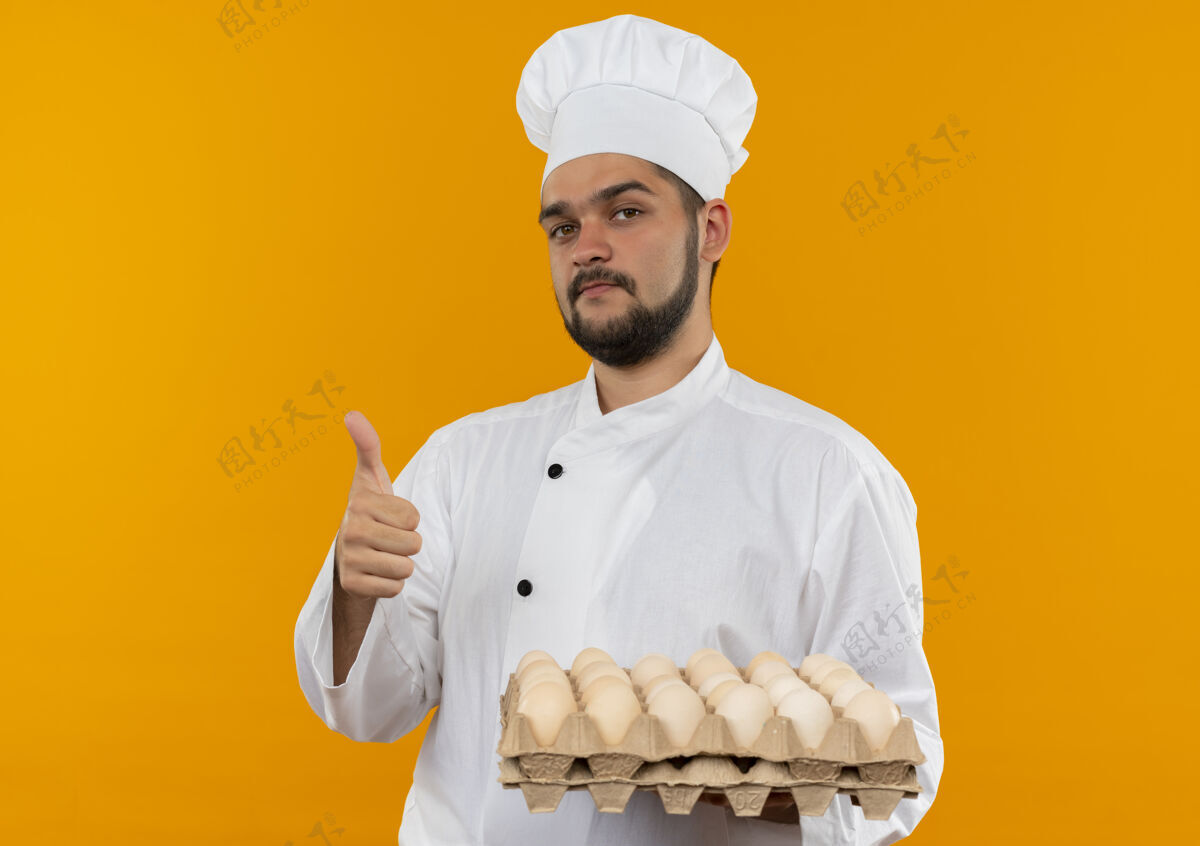 制服自信的年轻男性厨师身着厨师制服 手拿一盒鸡蛋 在橙色的墙上孤立地竖起大拇指信心年轻持有