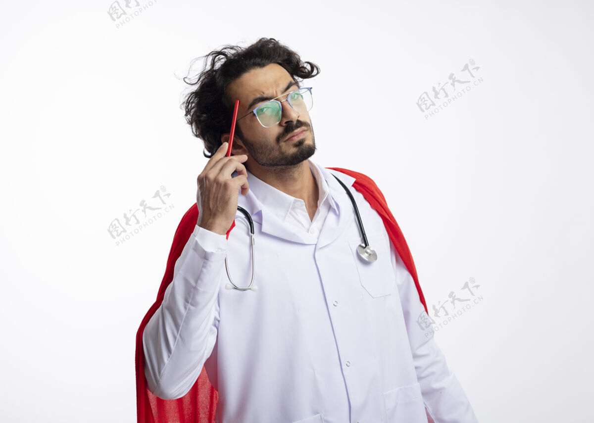 思想体贴的年轻白人超级英雄戴着眼镜 身穿医生制服 披着红色斗篷 脖子上戴着听诊器 把铅笔放在太阳穴上年轻复制铅笔