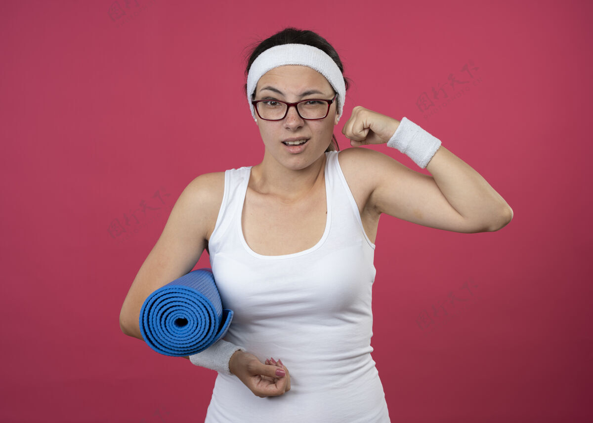 恼怒恼怒的年轻运动女孩戴着眼镜戴着头带和腕带紧张的二头肌 拿着运动垫空间眼镜光学