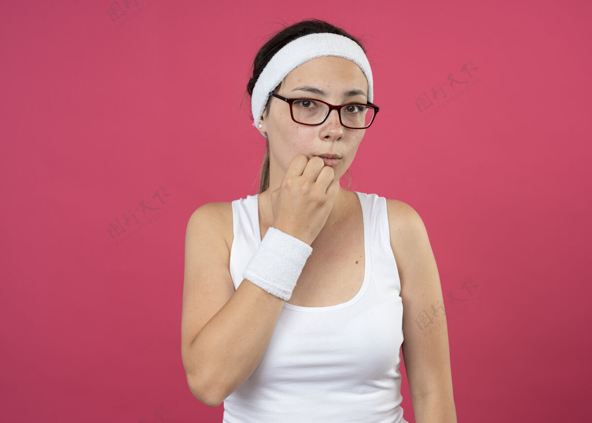 戴戴着眼镜 戴着头带和腕带的焦虑的年轻运动女孩把拳头放在下巴上粉色运动腕带