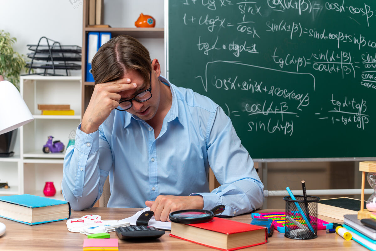 累年轻的男老师戴着眼镜坐在课桌旁 手里拿着书和笔记 在教室的黑板前又累又无聊书桌老师数学