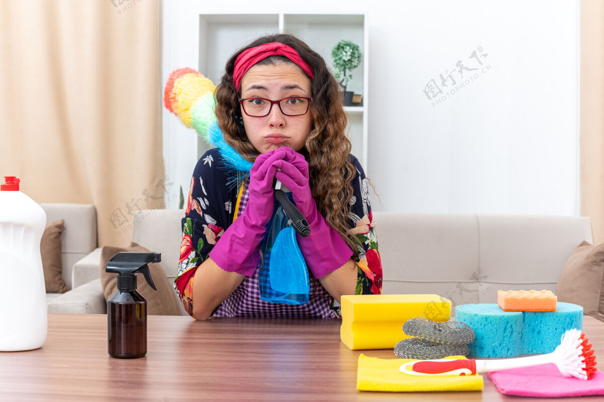 橡胶年轻女子手里拿着橡胶清洁喷雾剂和静电掸子 困惑地看着手套坐在桌子旁 手里拿着清洁用品和工具喷雾女人拿着
