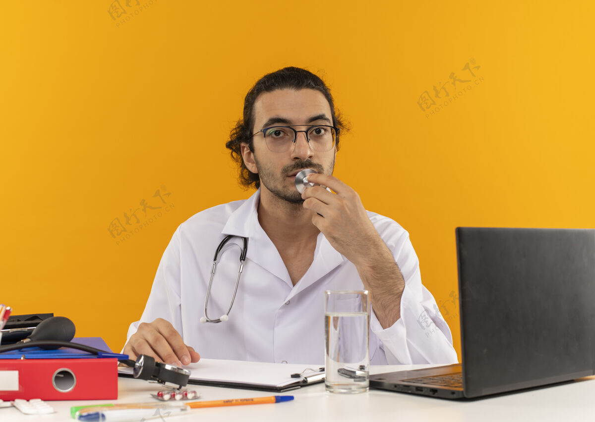 工具年轻的男医生戴着医用眼镜 穿着医用长袍 听诊器坐在桌子旁办公桌黄色年轻人