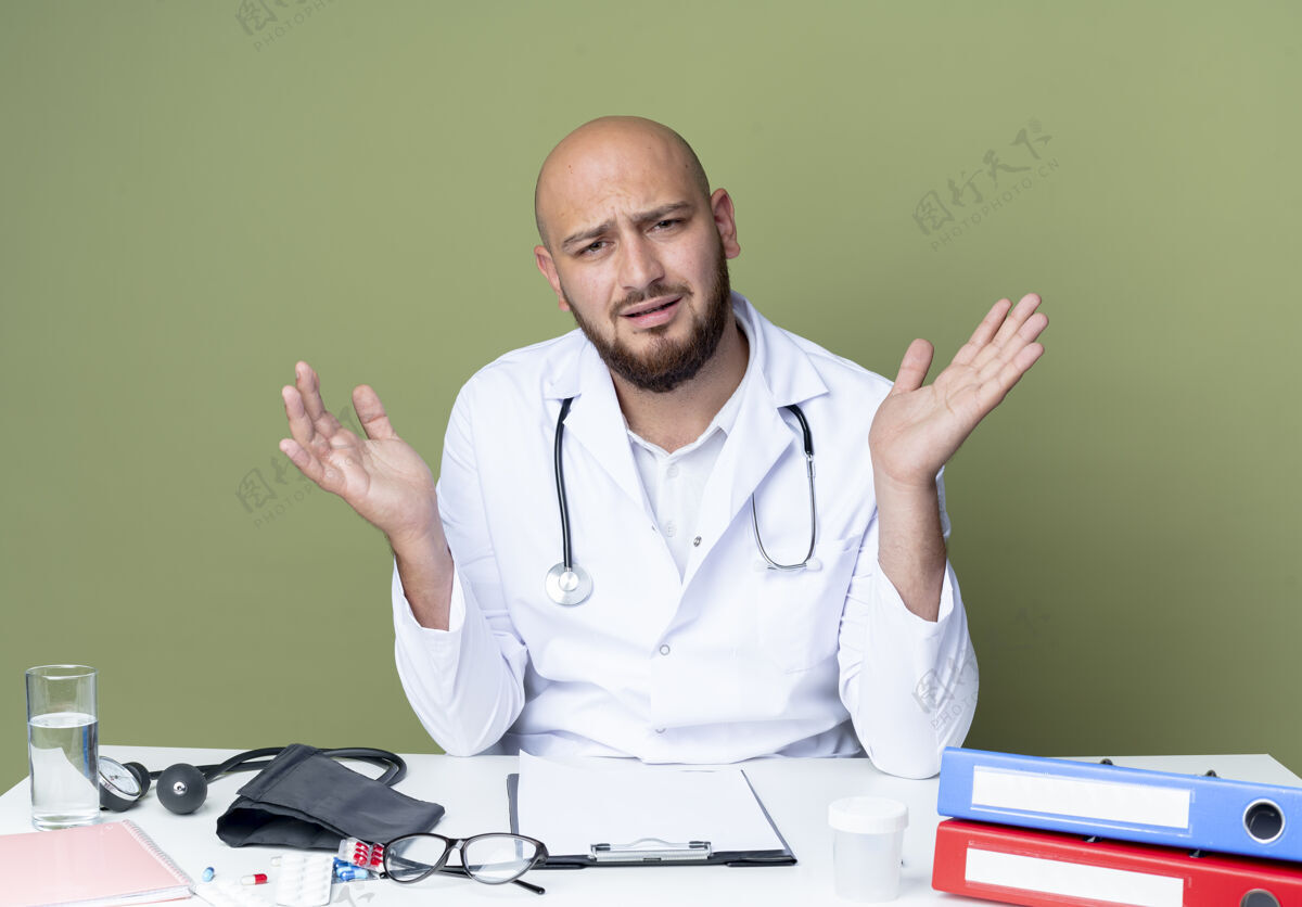 穿困惑的年轻秃头男医生穿着医用长袍和听诊器坐在办公桌旁听诊器蔓延坐着