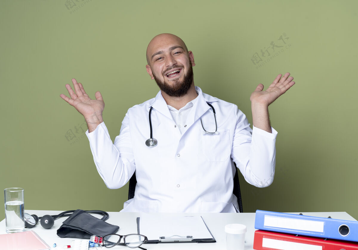 办公桌快乐的年轻秃头男医生穿着医用长袍和听诊器坐在办公桌旁秃头听诊器医疗