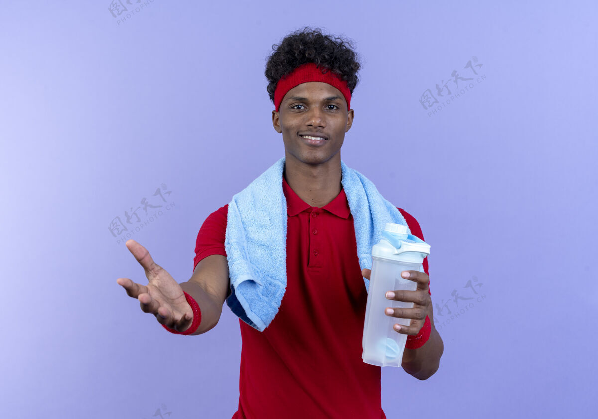 男人高兴的年轻黑人美国运动男子戴着头带和手镯拿着水瓶伸出手来相机与毛巾在肩上非裔美国人运动手握