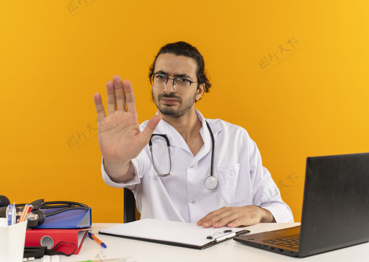 工作严格的年轻男性医生 戴着医用眼镜 穿着医用长袍 听诊器坐在办公桌旁黄色穿办公桌