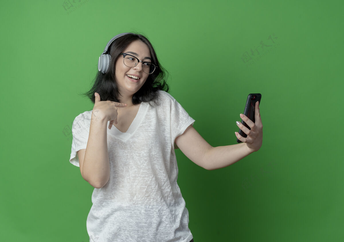 她自己快乐的年轻漂亮的白人女孩戴着眼镜和耳机拿着手机看着自己耳机穿女孩